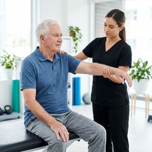 A senior man with white hair participates in a gentle physical therapy session guided by a female therapist in a bright, modern clinic.